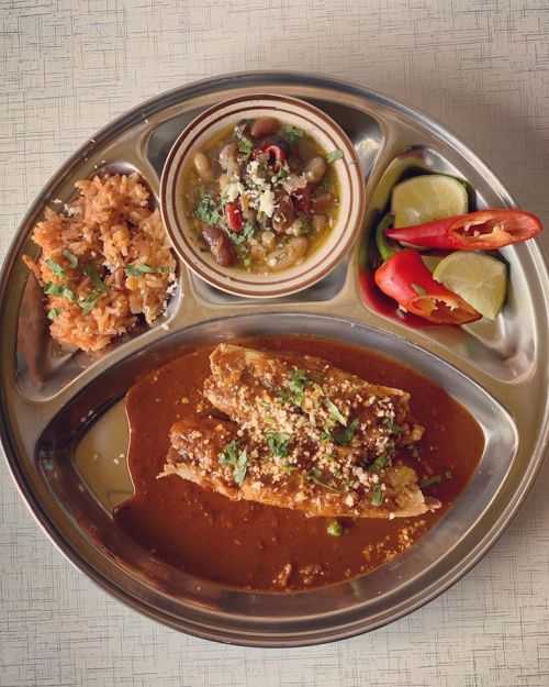 A silver platter with rice, beans, enchiladas in sauce, and fresh vegetables with lime wedges.