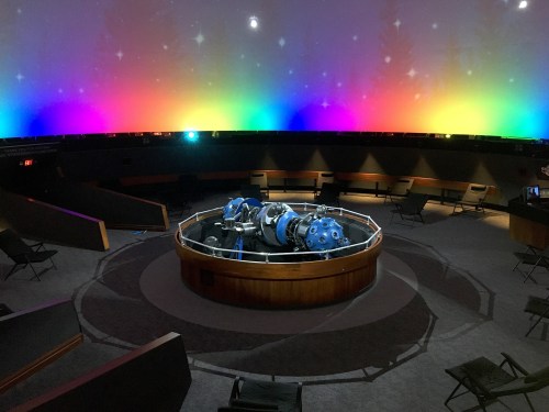 A planetarium interior featuring a star projector at the center, surrounded by colorful lights and seating.