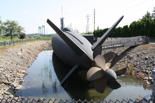A submarine with a large propeller is partially submerged in shallow water, surrounded by rocks and greenery.