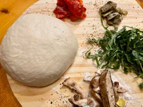 A wooden board with pizza dough, arugula, mushrooms, roasted peppers, and artichokes arranged around it.