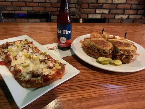 A plate with a cheesy flatbread topped with meat and veggies, alongside a sandwich and a bottle of beer on a wooden table.