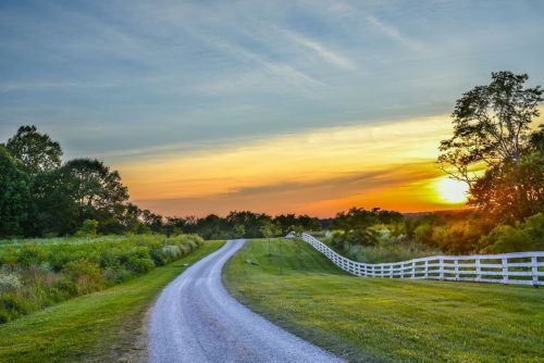 A winding gravel road leads through lush greenery, with a vibrant sunset illuminating the sky in the background.