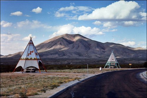 Two colorful tipis stand by a road, with a mountainous landscape and blue sky in the background.