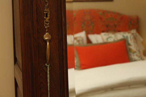 A close-up of a wooden cabinet handle with a blurred, cozy bedroom in the background featuring an orange headboard.