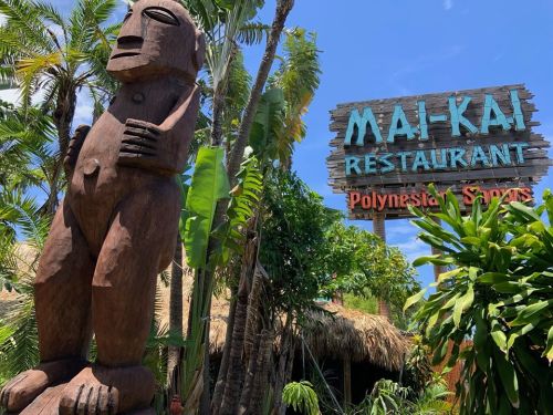 A wooden tiki statue stands in front of the Mai-Kai Restaurant sign, surrounded by tropical plants and blue skies.