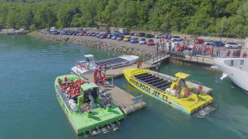 Two colorful boats docked at a marina, with people in life jackets preparing for a ride, surrounded by parked cars and greenery.