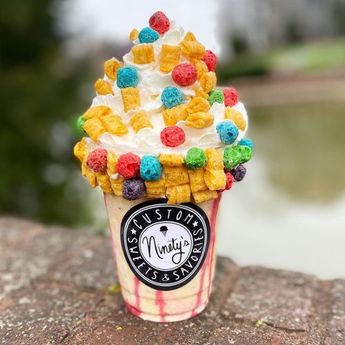 A colorful ice cream sundae topped with whipped cream and various cereal pieces, served in a branded cup.