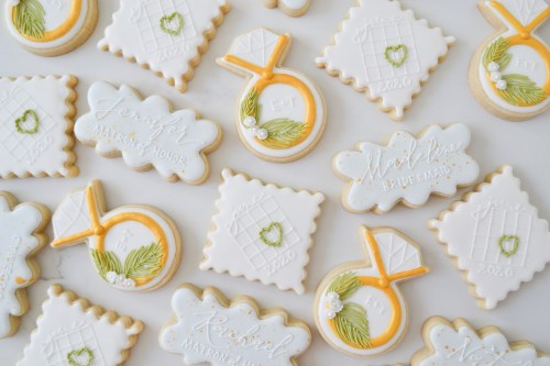 Decorative cookies featuring wedding themes, including rings and names, in white and gold icing on a light background.