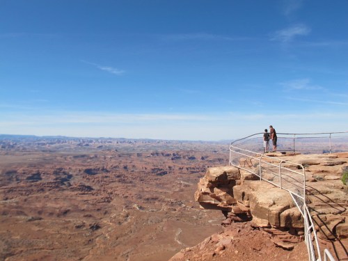 Two people stand on a rocky ledge with a railing, overlooking a vast desert landscape under a clear blue sky.