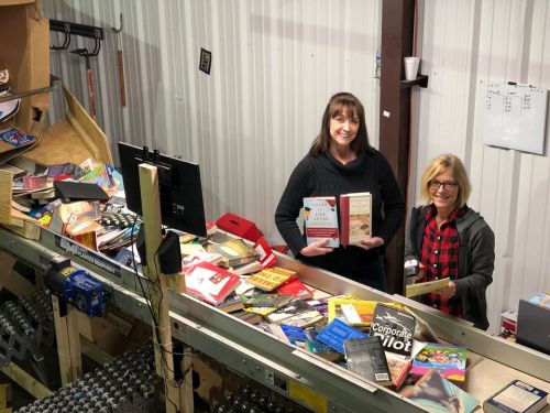 Two women stand behind a cluttered table filled with books, smiling and holding up a few titles in a warehouse setting.