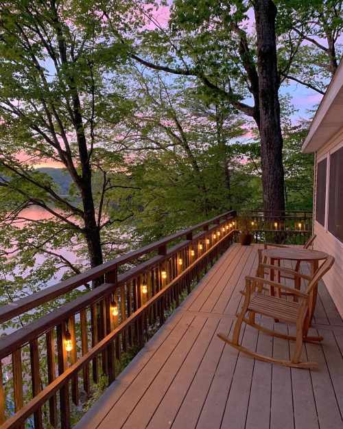 A serene lakeside deck at sunset, featuring two rocking chairs and string lights among lush green trees.