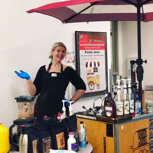 A smiling barista in an apron stands behind a coffee cart, ready to serve drinks under a red umbrella.
