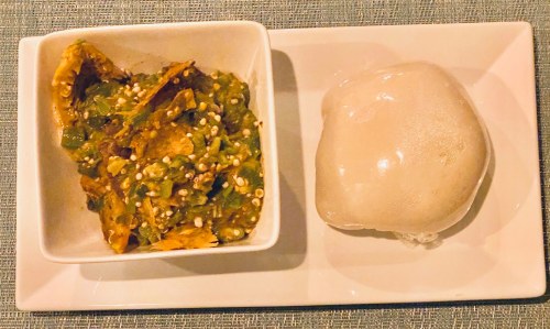 A plate with a serving of green vegetable stew topped with sesame seeds and a side of soft, round dough.