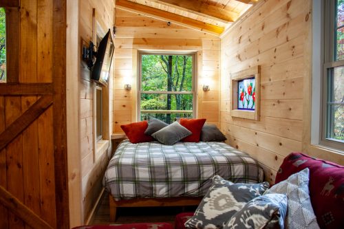 Cozy wooden cabin interior with a bed, colorful pillows, and large windows overlooking a forested area.