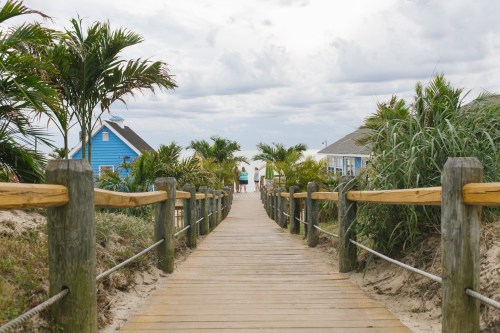 A wooden walkway leads through lush greenery to colorful beach houses under a cloudy sky.