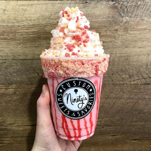 A colorful milkshake topped with whipped cream and sprinkles, served in a branded cup against a wooden background.