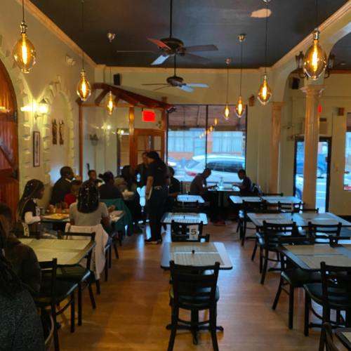 A cozy restaurant interior with wooden tables, chairs, and warm lighting, featuring diners and a server attending to guests.