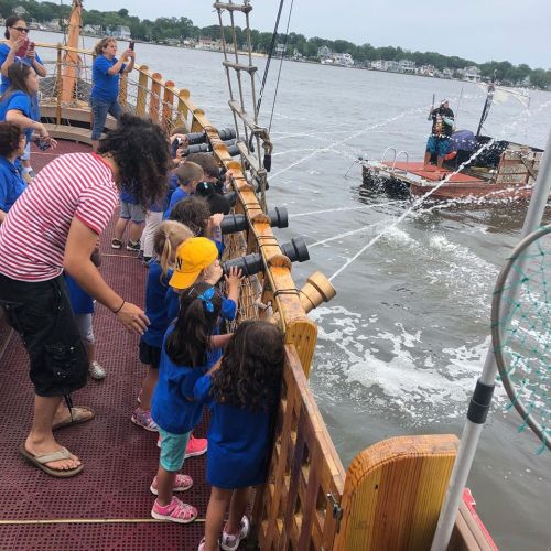 Children in blue shirts aim water cannons from a wooden ship, while another boat sprays water in the background.