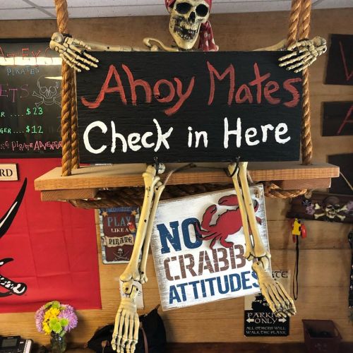 A skeleton dressed as a pirate holds a sign reading "Ahoy Mates, Check in Here" in a themed decor setting.