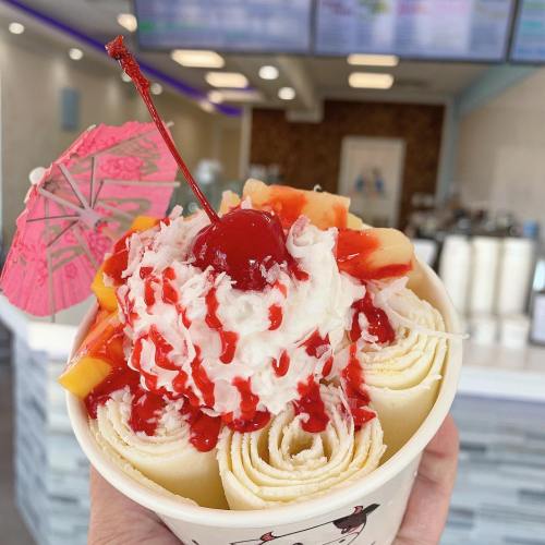 A hand holding a cup of rolled ice cream topped with whipped cream, fruit, cherry, and a pink umbrella.