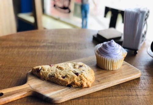 A wooden board with a cookie and a cupcake with purple frosting, set on a table in a cozy café.