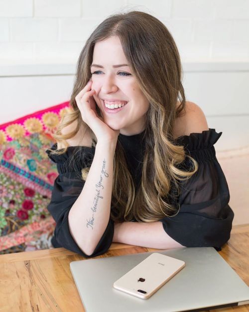 A woman with long hair smiles while resting her chin on her hand, sitting at a table with a laptop and phone.