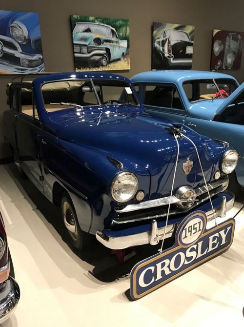 A vintage blue 1951 Crosley car displayed in a museum, with classic car paintings in the background.