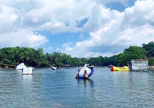 Colorful inflatable water toys float on a lake under a partly cloudy sky, surrounded by lush green trees.