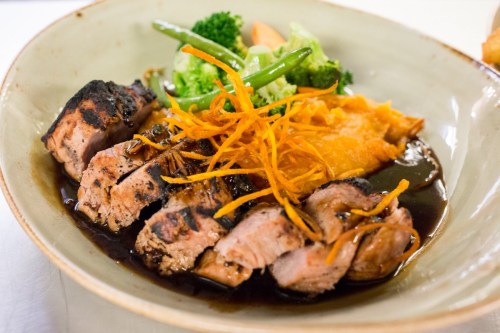 A plate of sliced grilled meat with a rich sauce, garnished with orange strips, served with vegetables and mashed sweet potato.