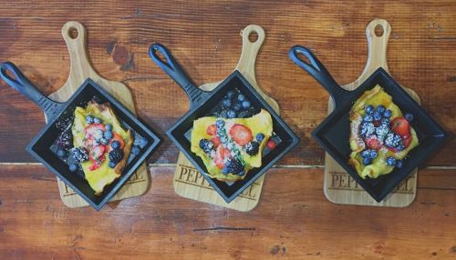 Three small cast iron pans on wooden boards, each filled with colorful omelets topped with berries and powdered sugar.