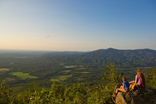 Two hikers sit on a rock, overlooking a vast green valley and distant mountains under a clear sky.
