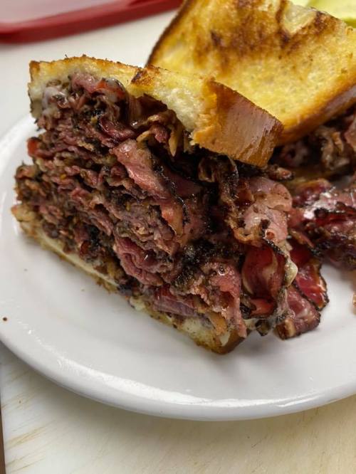 A close-up of a stacked pastrami sandwich on toasted bread, served on a white plate.