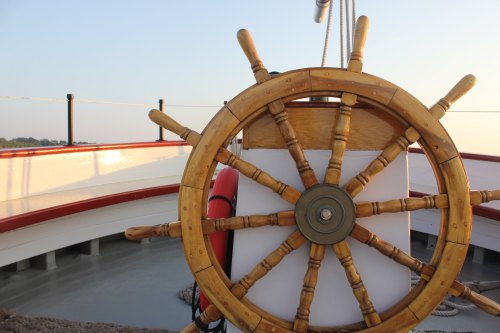 A wooden ship's wheel stands prominently on a boat, with a serene sunset in the background.