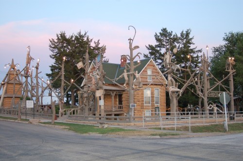 A whimsical wooden structure with sculptures and lights, surrounded by trees, set against a pastel sky at dusk.