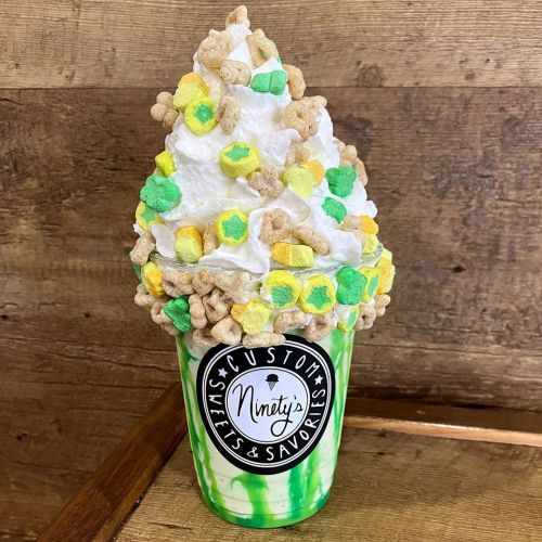 A colorful milkshake topped with whipped cream, cereal, and marshmallows in a clear cup with a logo.