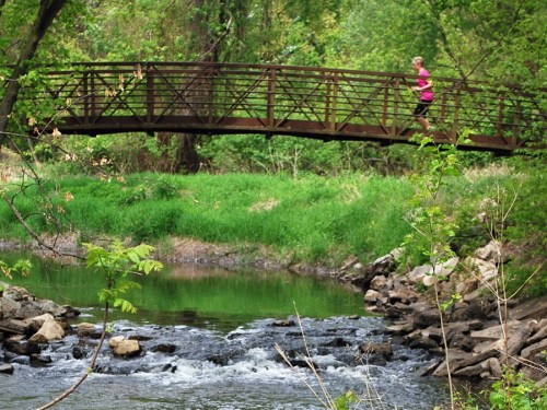A person jogs across a wooden bridge over a stream, surrounded by lush greenery and rocks.
