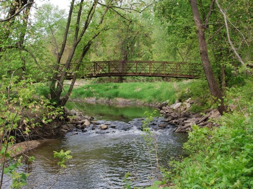 A serene river scene with a wooden bridge over water, surrounded by lush green trees and rocky banks.
