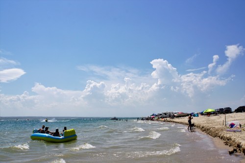 A sunny beach scene with people in a boat, sandy shore, and fluffy clouds in a blue sky.