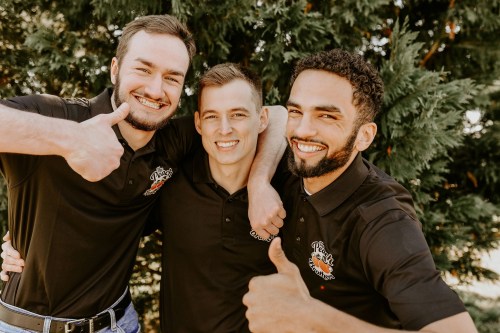 Three smiling men in matching black polo shirts give thumbs up in front of a green tree backdrop.