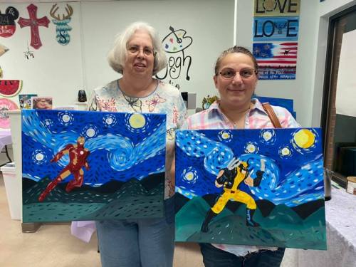 Two women hold up colorful paintings of superhero characters against a starry night background.