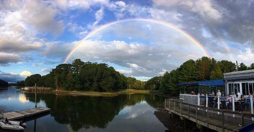 A vibrant rainbow arches over a calm lake, reflecting trees and a restaurant patio with diners.