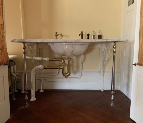 A vintage sink with a marble top and brass fixtures, supported by decorative metal legs, in a room with wooden flooring.