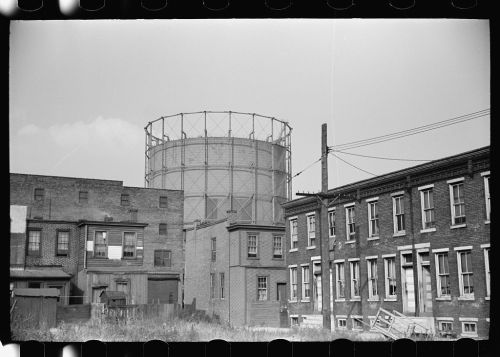 Black and white photo of a gas storage tank behind old brick buildings and overgrown grass.