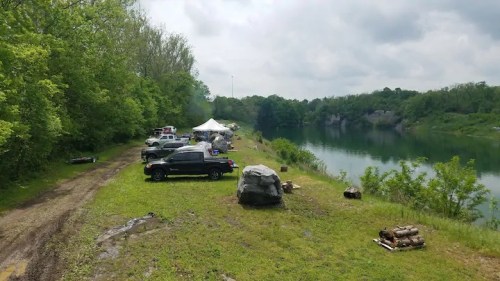 A scenic lakeside campsite with parked vehicles, tents, and lush greenery under a cloudy sky.