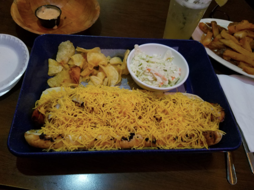 A plate of food featuring a large cheese-covered dish, chips, coleslaw, and a drink on the side.
