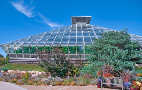 A modern glass greenhouse surrounded by colorful plants and a bench under a clear blue sky.