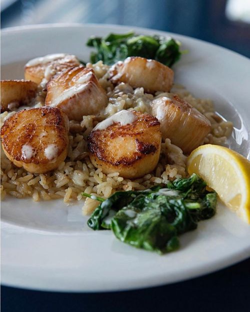 A plate of seared scallops on rice, accompanied by sautéed spinach and a lemon wedge.