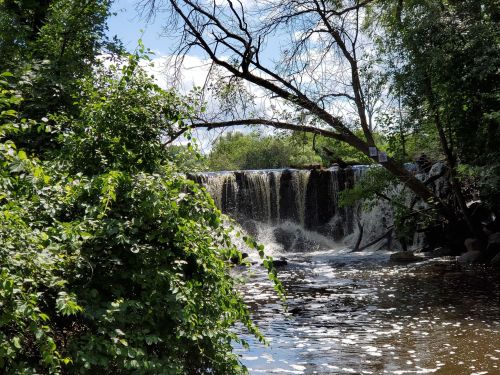 A serene waterfall cascades over rocks, surrounded by lush greenery and trees under a bright blue sky.