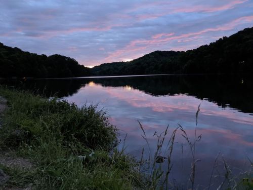 A serene lake at dusk, reflecting colorful clouds and surrounded by lush greenery and hills.