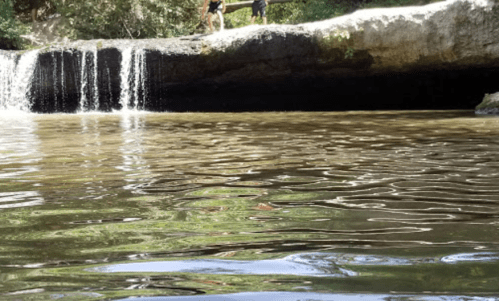 Two people stand on a rocky ledge near a waterfall, with water cascading into a calm pool below.